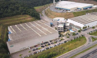 A imagem mostra uma vista aérea de um complexo industrial cercado por áreas verdes e uma estrada.