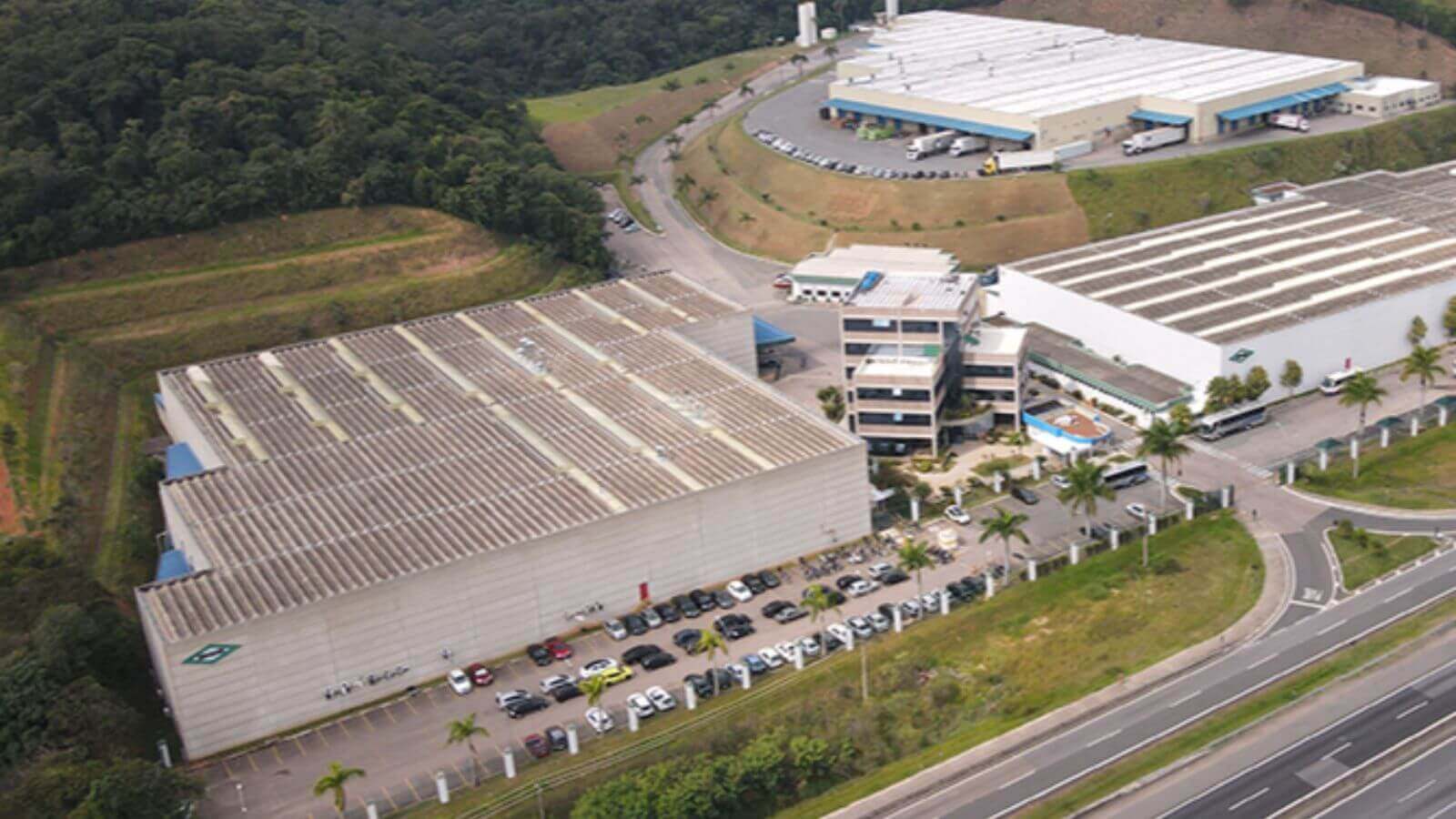 A imagem mostra uma vista aérea de um complexo industrial cercado por áreas verdes e uma estrada.
