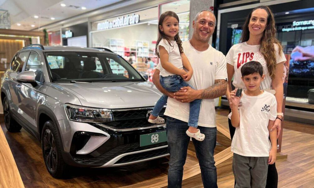 A imagem mostra uma família de quatro pessoas posando ao lado de um carro novo em um shopping. O grupo inclui um homem, uma mulher, uma menina e um menino.