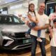 A imagem mostra uma família de quatro pessoas posando ao lado de um carro novo em um shopping. O grupo inclui um homem, uma mulher, uma menina e um menino.