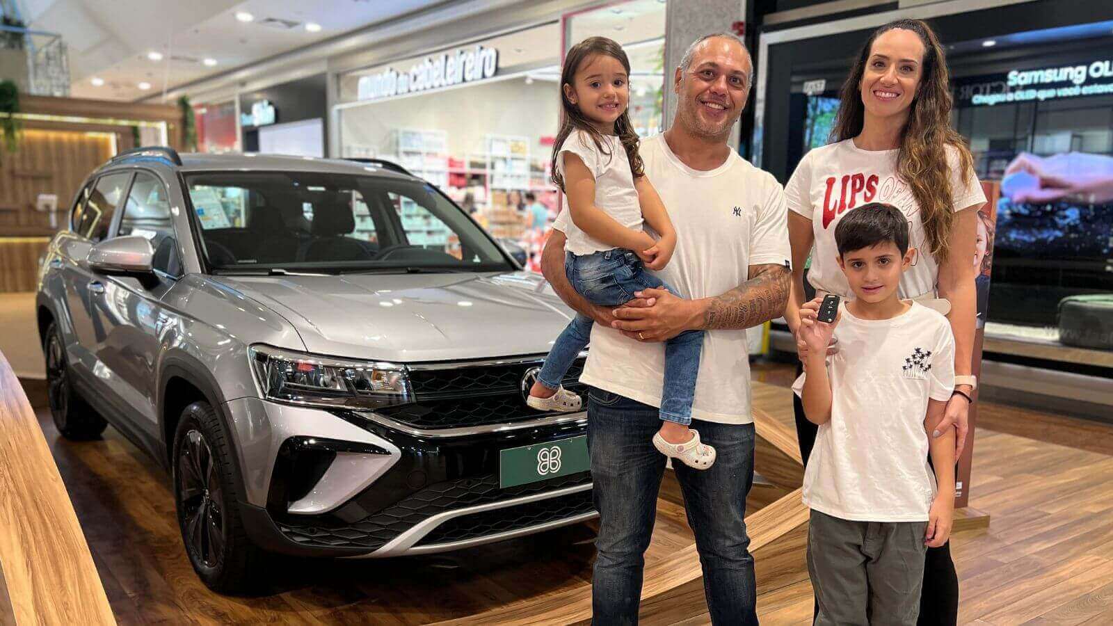 A imagem mostra uma família de quatro pessoas posando ao lado de um carro novo em um shopping. O grupo inclui um homem, uma mulher, uma menina e um menino.
