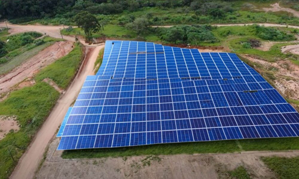 Na foto, vemos uma usina fotovoltaica em construção. A imagem mostra uma grande área coberta por inúmeras placas solares organizadas de maneira ordenada. O local onde a usina está situada é uma região verde e aberta, com vegetação ao redor e algumas estradas de terra que circundam o terreno. As placas solares estão dispostas em linhas e colunas, ocupando um espaço considerável, indicando um projeto de grande escala. A usina está localizada em uma área rural, longe de grandes edificações, o que permite um aproveitamento eficiente da luz solar.