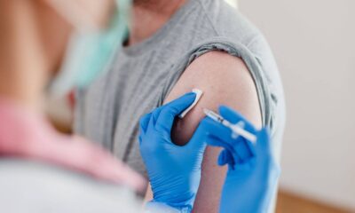 Pessoa de camiseta cinza recebendo vacina através de seringa no braço esquerdo