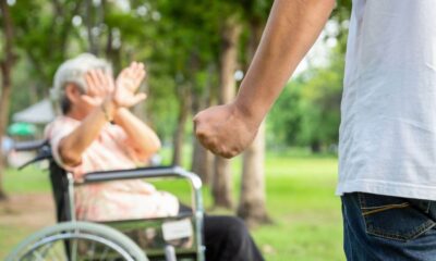 Imagem mostra uma idosa em cadeira de rodas sendo ameaçada por um homem com punho fechado em um parque, simbolizando violência contra idosos.