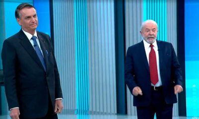 Políticos Jair Bolsonaro e Luiz Inácio Lula da Silva em debate na TV, em pé lado a lado, com fundo azul de estúdio.