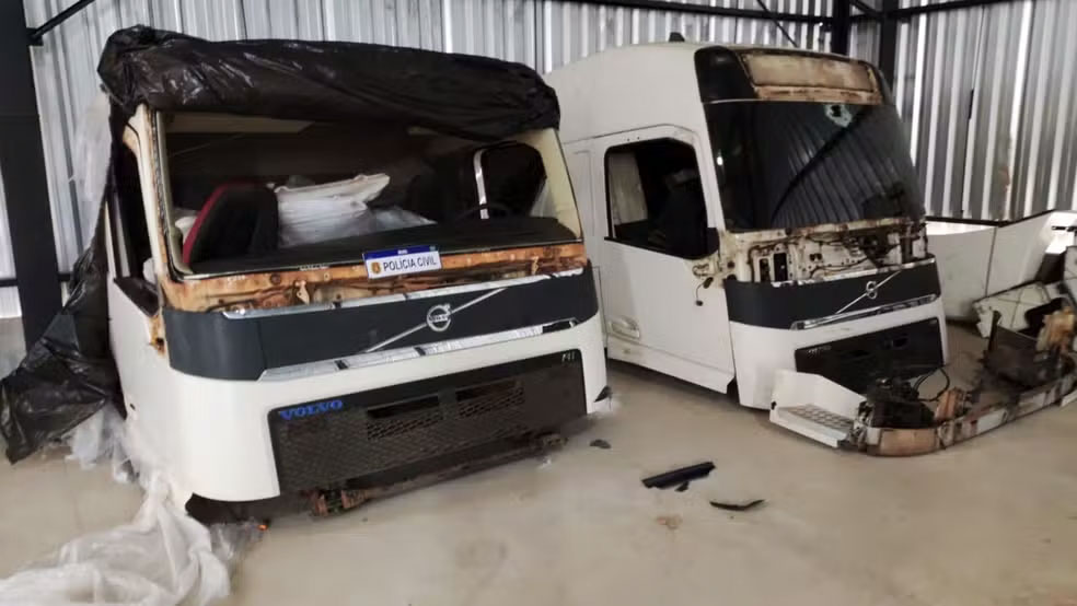 Duas cabines de caminhão Volvo danificadas, sem partes frontais e em ambiente coberto, com placa da Polícia Civil visível.