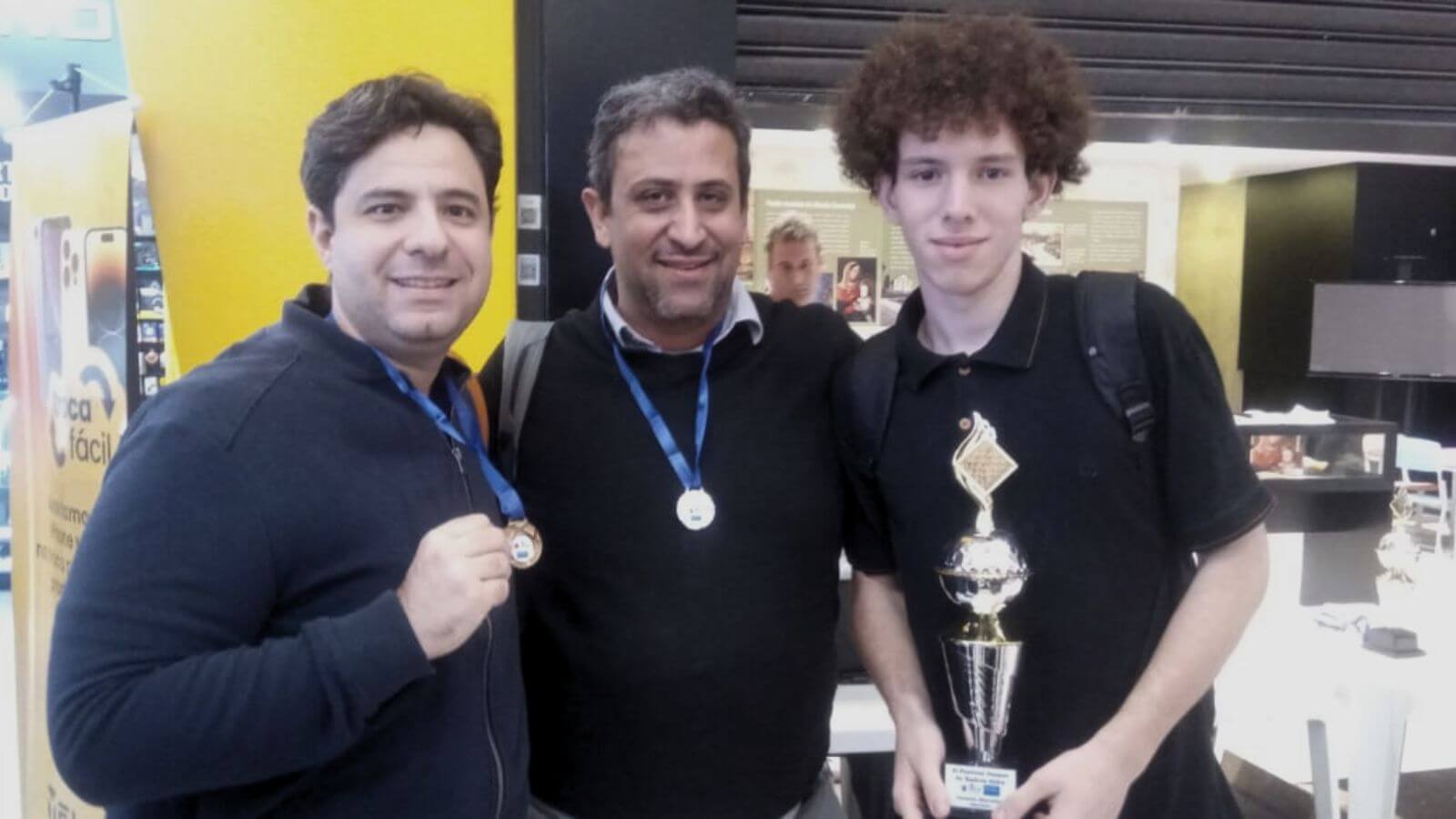 Três homens do Time Jundiaí de Xadrez posando com medalhas e um troféu, celebrando uma conquista em um ambiente de competição.