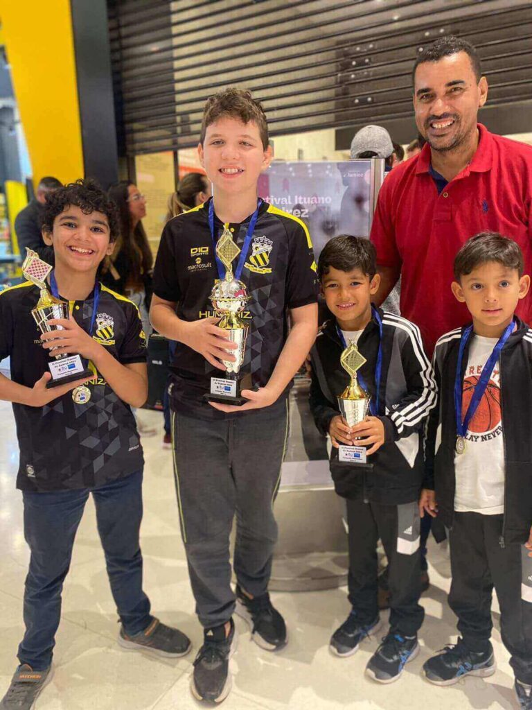 Grupo de crianças e um adulto sorrindo com troféus e medalhas, comemorando uma vitória em um ambiente de competição.