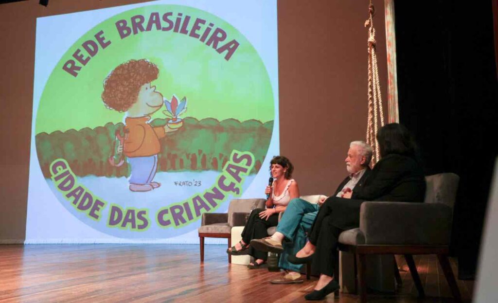 três pessoas em um painel discutindo a "Rede Brasileira Cidade das Crianças", com um fundo de uma ilustração infantil.