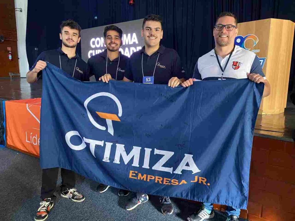 Quatro homens sorridentes seguram uma bandeira da Otimiza Empresa Jr. durante um evento, com um palco ao fundo.