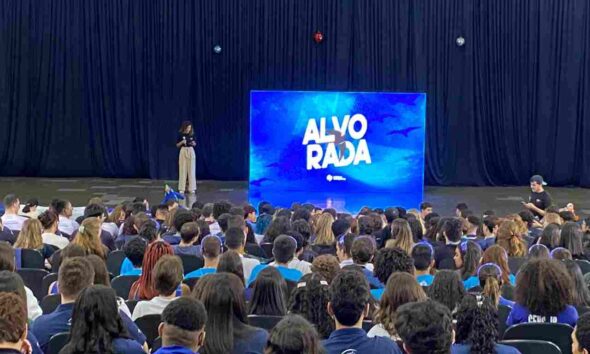Palestrante se apresenta em um auditório cheio de pessoas, com uma tela grande ao fundo exibindo "Alvorada" em azul.