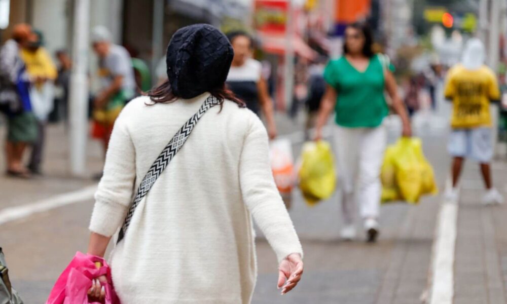 Mulher com touca preta e casaco branco caminha em rua movimentada de Jundiaí em dia frio, segurando sacola rosa.