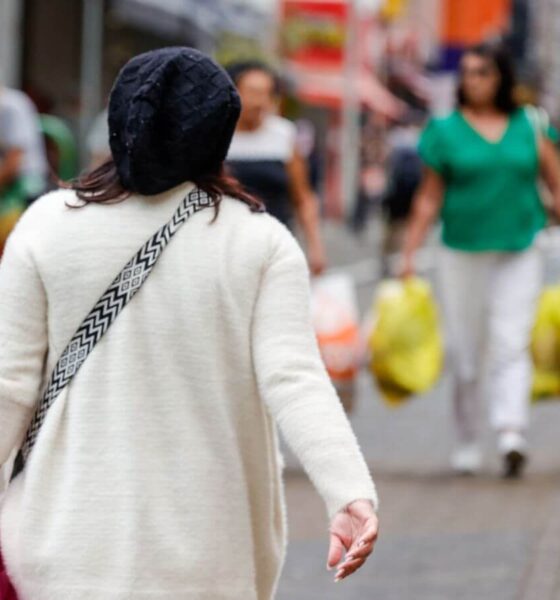 Mulher com touca preta e casaco branco caminha em rua movimentada de Jundiaí em dia frio, segurando sacola rosa.