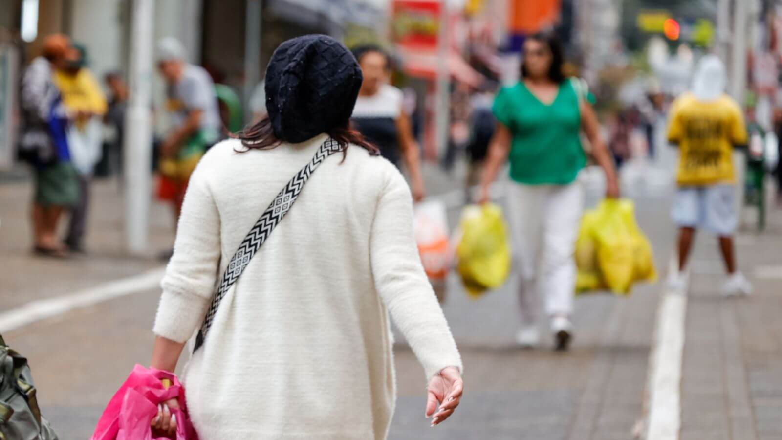 Mulher com touca preta e casaco branco caminha em rua movimentada de Jundiaí em dia frio, segurando sacola rosa.