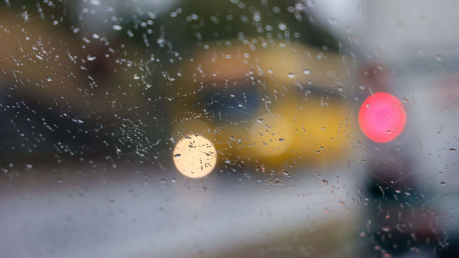 Gotas de chuva embaçam vidro, destacando luzes desfocadas de carros na rua em dia chuvoso em Jundiaí.