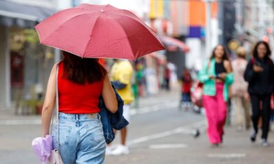 Mulher com guarda-chuva vermelho caminha em rua movimentada de Jundiaí em dia chuvoso, vestindo blusa vermelha e jeans em vias de Jundiaí.