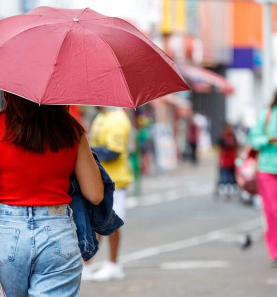 Mulher com guarda-chuva vermelho caminha em rua movimentada de Jundiaí em dia chuvoso, vestindo blusa vermelha e jeans em vias de Jundiaí, rementendo à previsão do tempo.