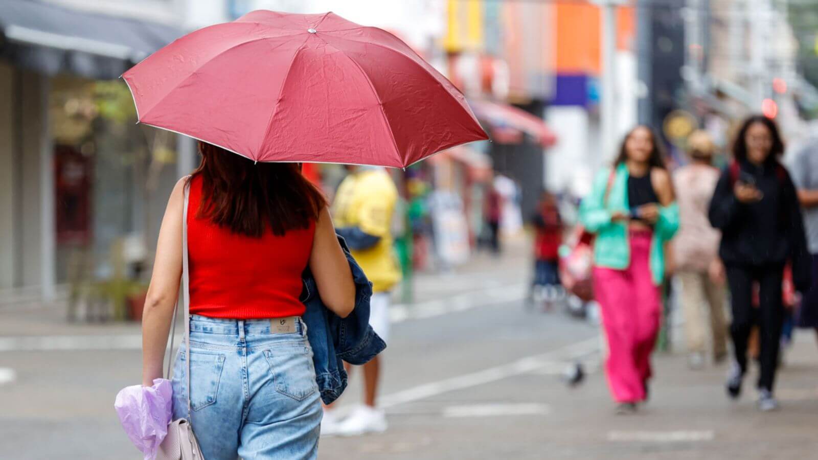 Mulher com guarda-chuva vermelho caminha em rua movimentada de Jundiaí em dia chuvoso, vestindo blusa vermelha e jeans em vias de Jundiaí.