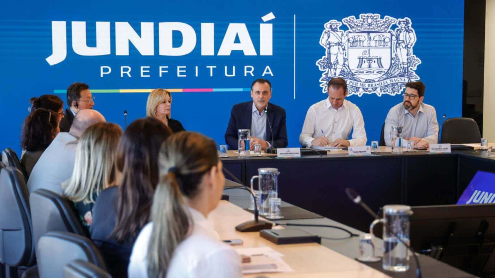 Autoridades de Jundiaí em reunião com representantes do Reino Unido sob um banner azul da prefeitura da cidade.