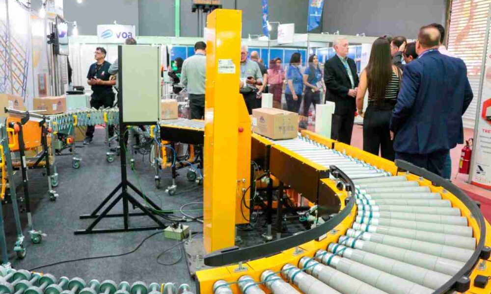 A imagem mostra uma exposição ou feira de tecnologia industrial, onde pessoas estão observando e discutindo equipamentos de automação, como transportadores de rolos usados para movimentação de caixas. No centro, há uma máquina amarela com esteiras rolantes que transportam caixas de papelão. À direita, um grupo de pessoas, algumas de terno, conversa próximo aos estandes.