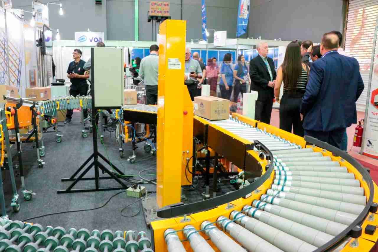 A imagem mostra uma exposição ou feira de tecnologia industrial, onde pessoas estão observando e discutindo equipamentos de automação, como transportadores de rolos usados para movimentação de caixas. No centro, há uma máquina amarela com esteiras rolantes que transportam caixas de papelão. À direita, um grupo de pessoas, algumas de terno, conversa próximo aos estandes.