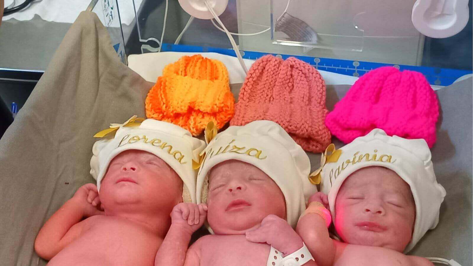 Trigêmeas recém-nascidas, Lorena, Luiza e Lavínia, deitadas lado a lado na maternidade do Hospital Universitário de Jundiaí com gorros coloridos.