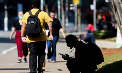 Pessoas caminhando em calçada urbana; um homem de mochila amarela em destaque; outro agachado, concentrado no celular.