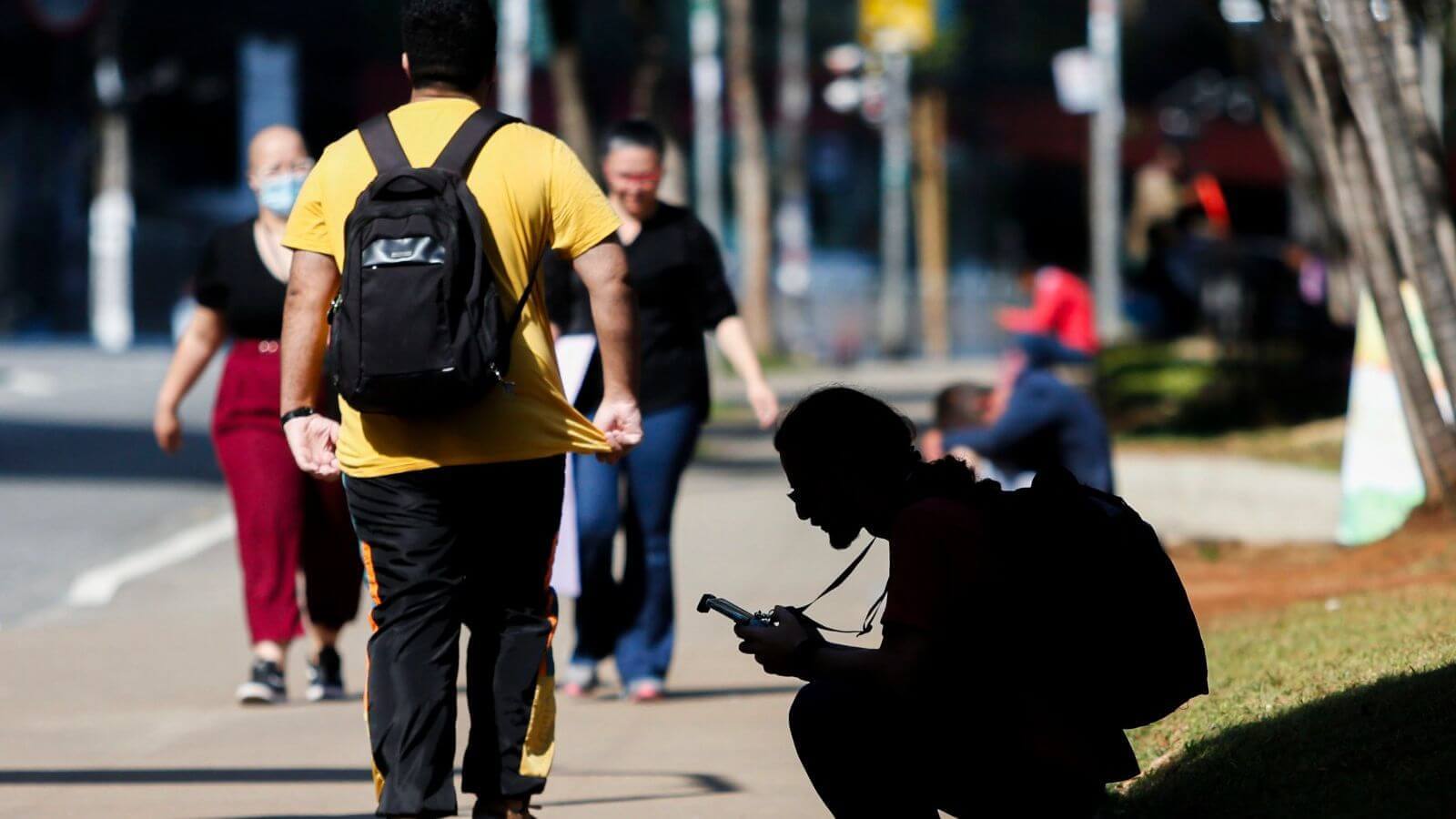 Pessoas caminhando em calçada urbana; um homem de mochila amarela em destaque; outro agachado, concentrado no celular.