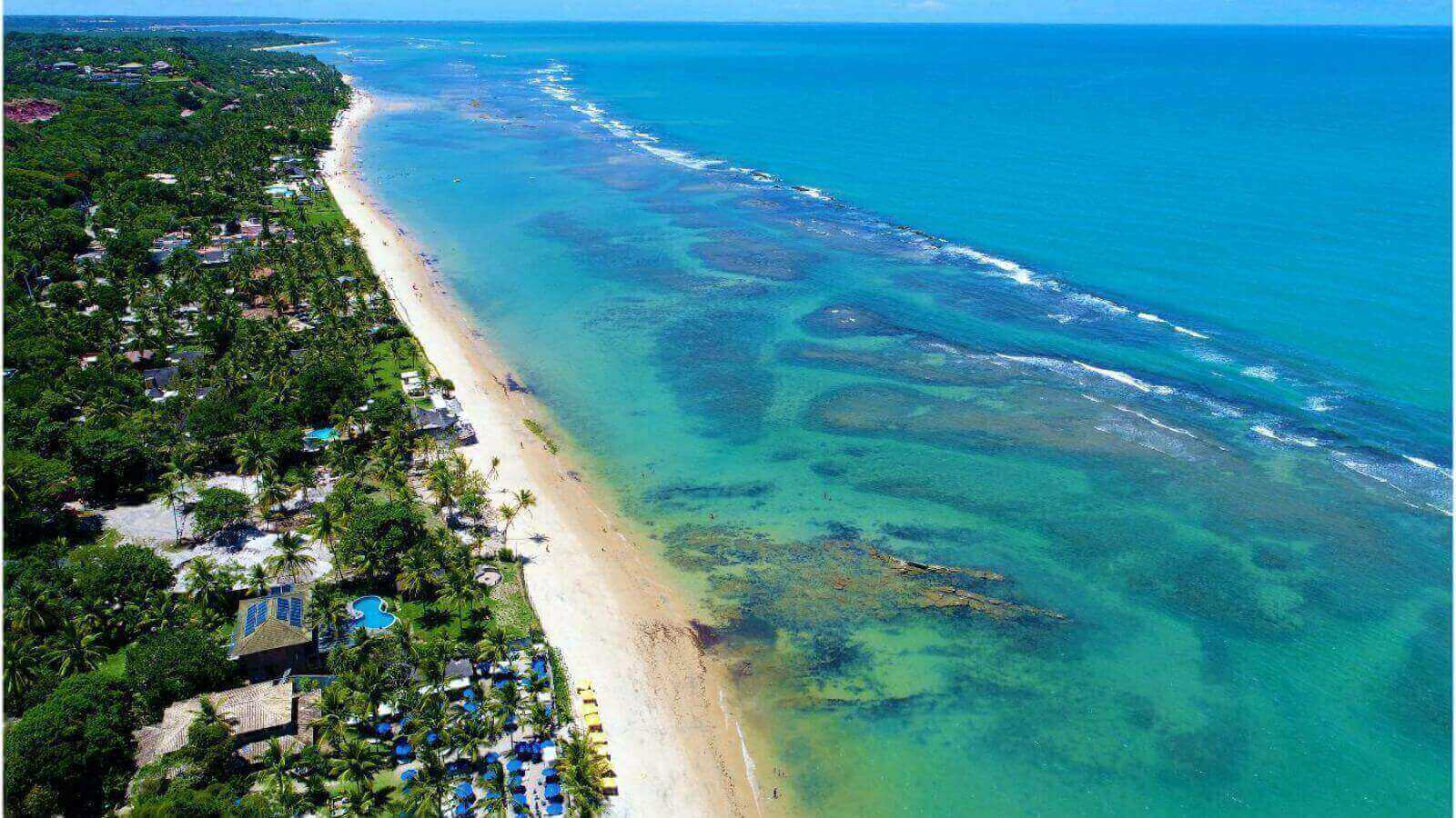Praia paradisíaca em Trancoso, Bahia, com águas cristalinas, faixa de areia extensa e vegetação exuberante ao longo da costa.