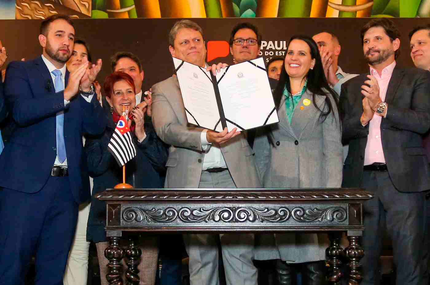 Governador Tarcísio de Freitas segurando e exibindo um documento assinado, rodeado por um grupo de pessoas aplaudindo