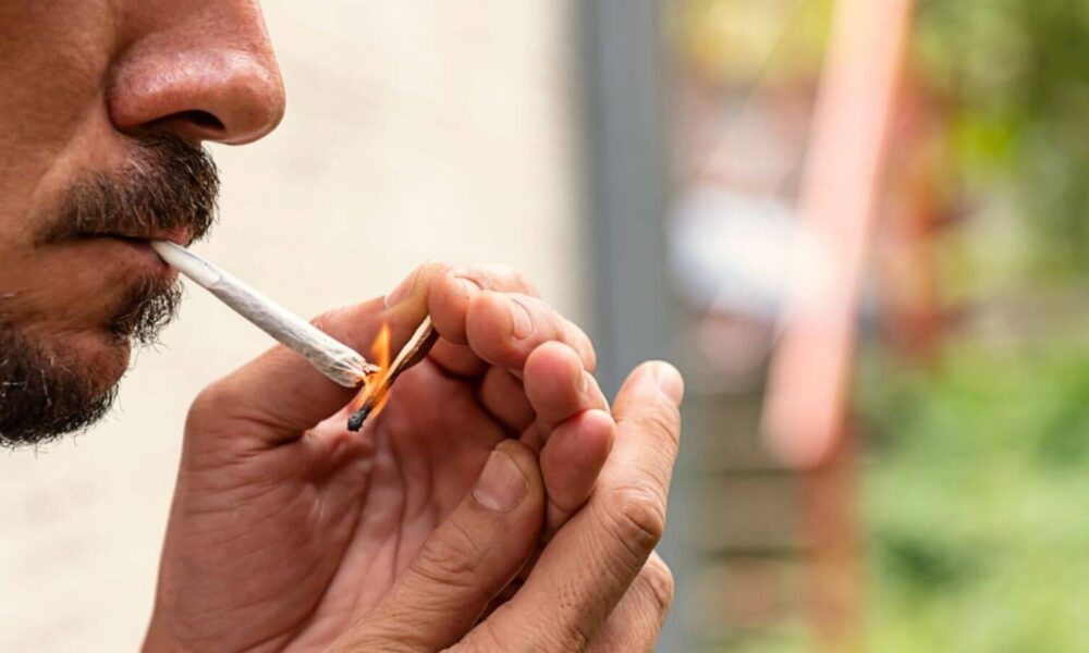 Homem acendendo um baseado de maconha ao ar livre, close-up no rosto e nas mãos, destacando o uso da substância.