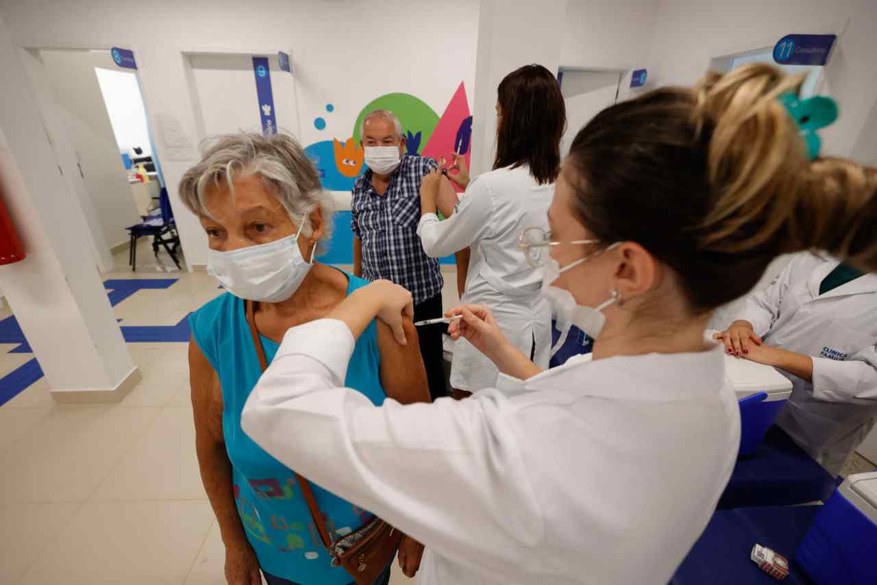 Mulher idosa recebe vacina contra COVID-19 em Jundiaí, com profissionais de saúde ao fundo.