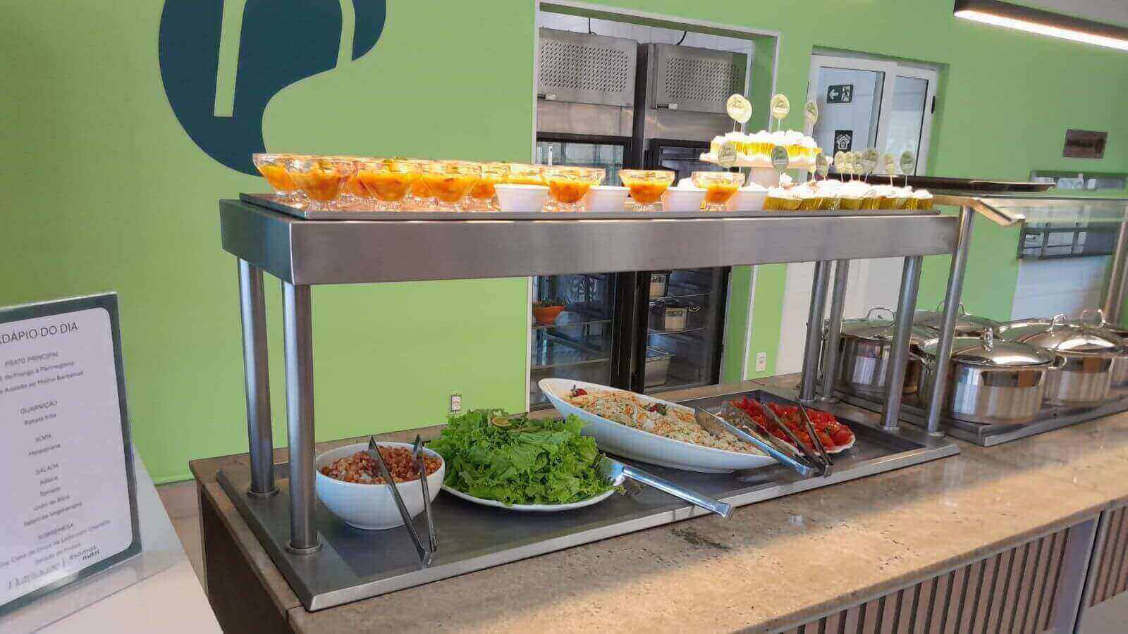 Buffet de saladas e sobremesas em restaurante, com cardápio do dia exposto, destacando ambiente moderno e organizado.