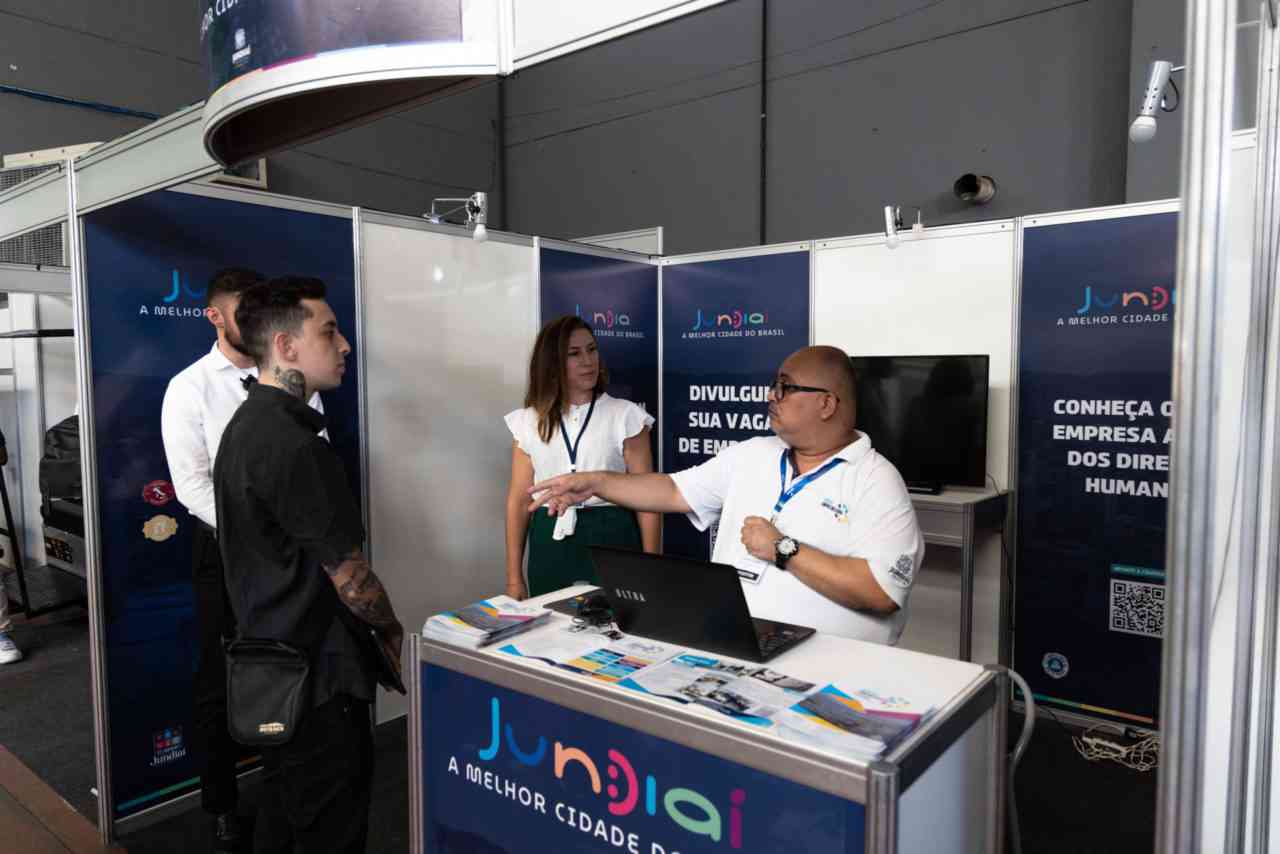 Stand da Prefeitura na Fatec Log 2024 na feira, com quatro pessoas conversando e banners de Jundiaí ao fundo, destacando oportunidades de emprego.