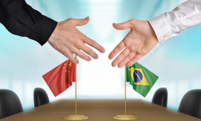 Dois homens iniciando um aperto de mãos com uma bandeira da China e uma bandeira do Brasil em cima de uma mesa