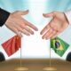 Dois homens iniciando um aperto de mãos com uma bandeira da China e uma bandeira do Brasil em cima de uma mesa