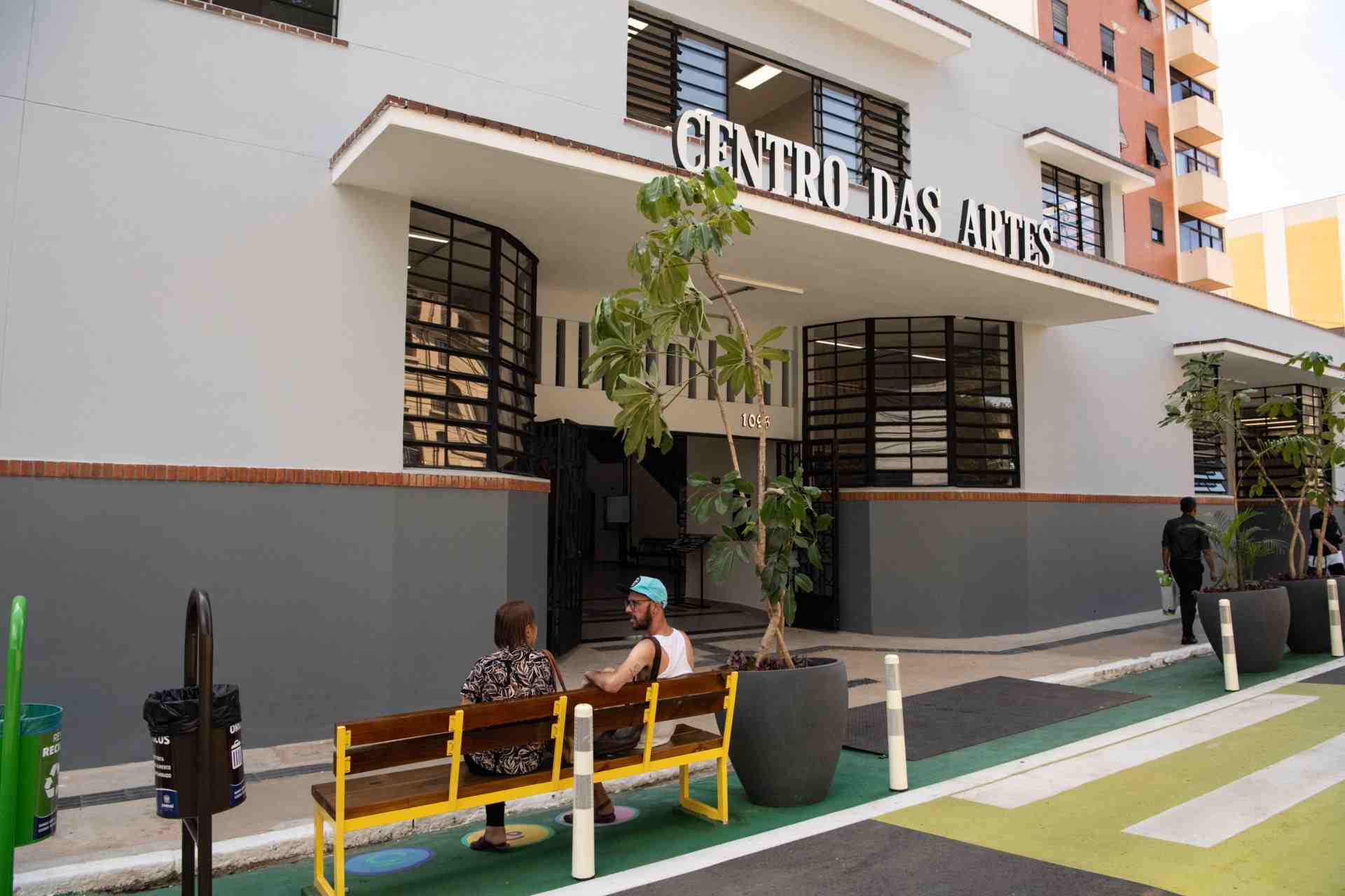 Entrada do Centro das Artes em Jundiaí, com duas pessoas sentadas em um banco na frente e plantas decorativas ao redor.