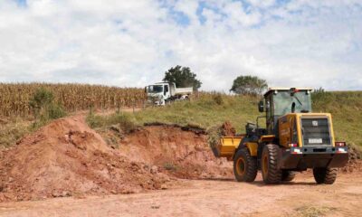 Trator realizando terraplanagem em uma área rural, com um caminhão ao fundo e uma plantação ao lado.