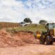 Trator realizando terraplanagem em uma área rural, com um caminhão ao fundo e uma plantação ao lado.