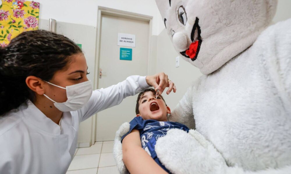 Criança deitada em colo de mascote do Zé Gotinha recebendo vacinação contra paralisia infantil