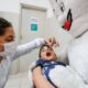 Criança deitada em colo de mascote do Zé Gotinha recebendo vacinação contra paralisia infantil