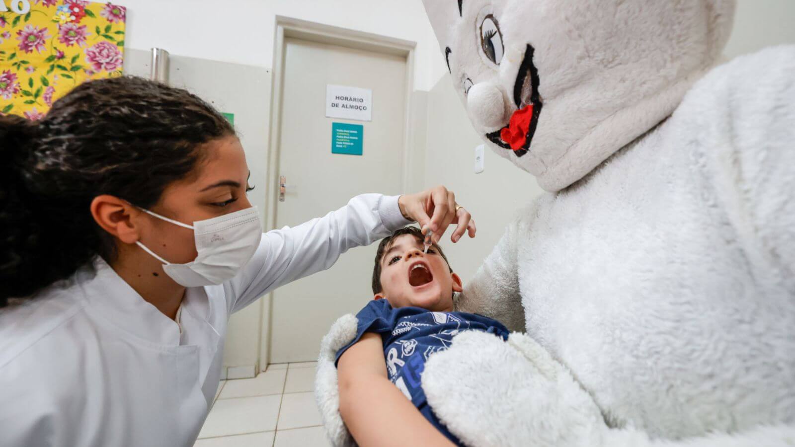 Criança deitada em colo de mascote do Zé Gotinha recebendo vacinação contra paralisia infantil