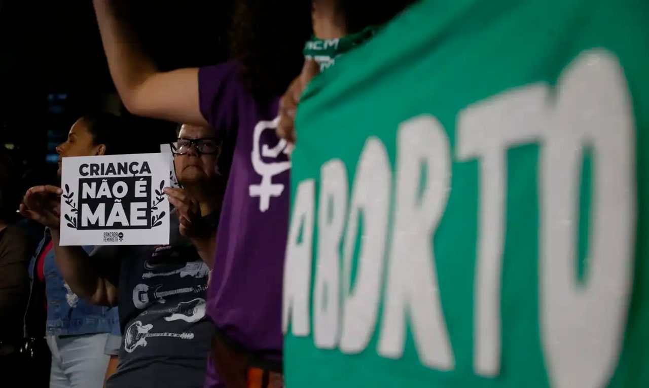 Protesto com placas sobre direitos reprodutivos e aborto; uma diz 'Criança não é mãe'. Legislação sobre aborto pelo mundo.