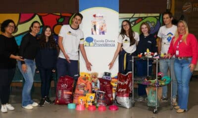 Adultos e jovens ao lado de um banner da Escola Divina Providência de Jundiaí. À frente deles, itens doados, incluindo sacos de ração para cães e gatos e brinquedos para animais.