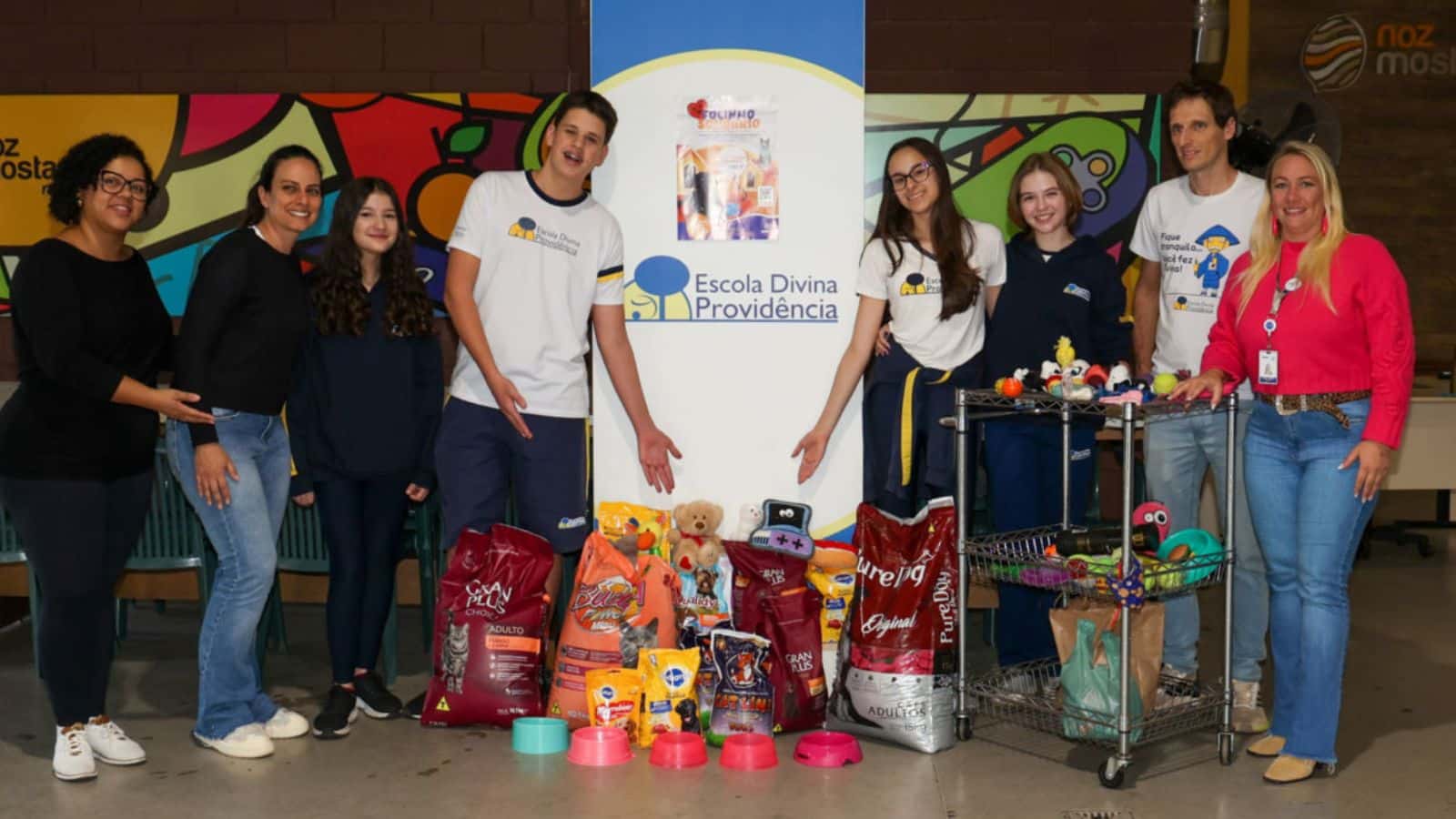 Adultos e jovens ao lado de um banner da Escola Divina Providência de Jundiaí. À frente deles, itens doados, incluindo sacos de ração para cães e gatos e brinquedos para animais.