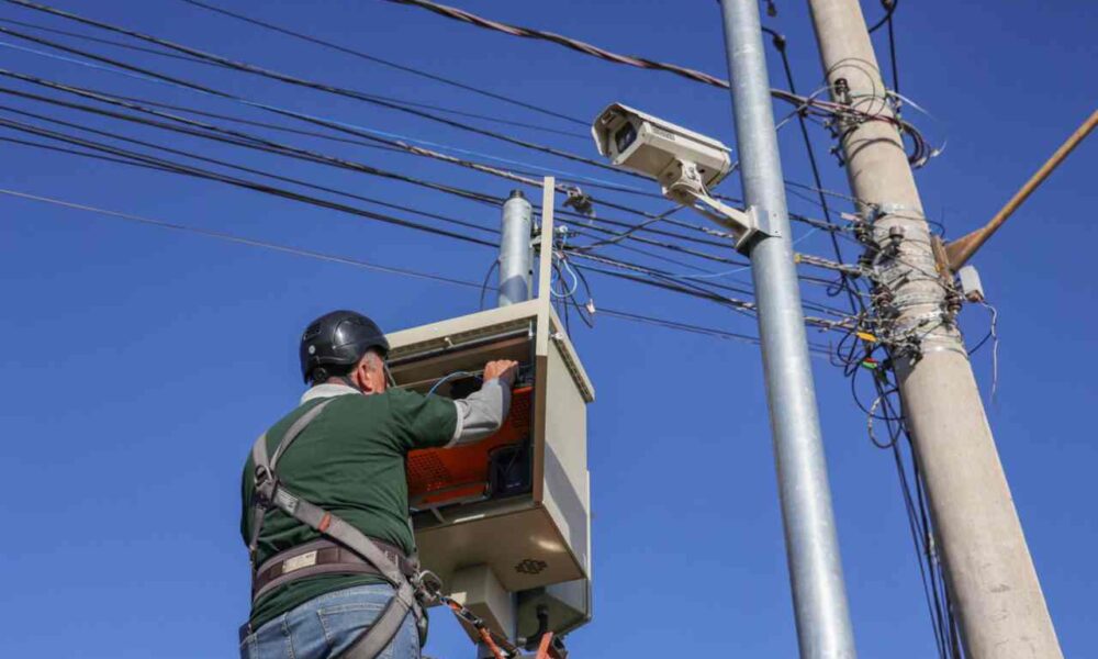 Homem instala câmeras de monitoramento em poste em Jundiaí, com fiação elétrica visível e céu azul ao fundo.