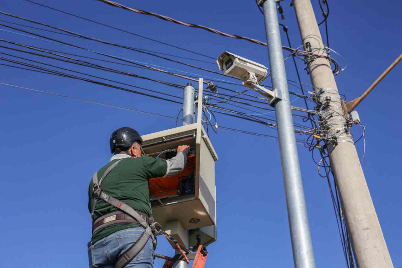 Homem instala câmeras de monitoramento em poste em Jundiaí, com fiação elétrica visível e céu azul ao fundo.