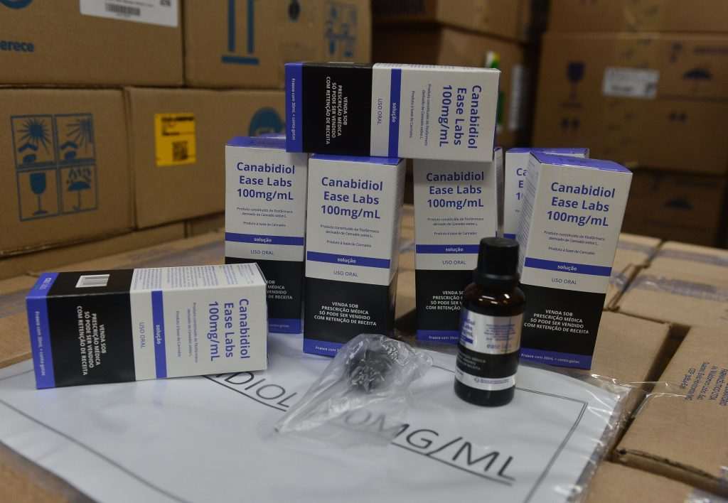 Caixas e frasco de Canabidiol Ease Labs 100mg/mL, empilhados em um depósito, indicando sua distribuição pelo SUS em São Paulo.