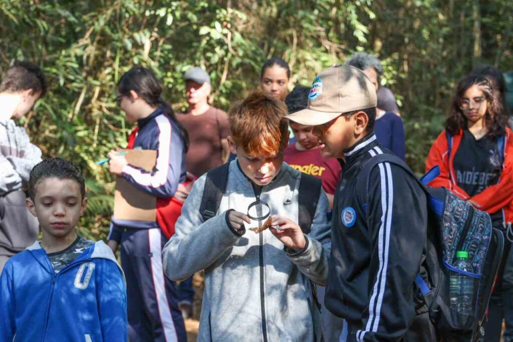Crianças e jovens observam insetos com uma lupa durante atividade educativa na Serra do Japi, rodeados por vegetação densa.