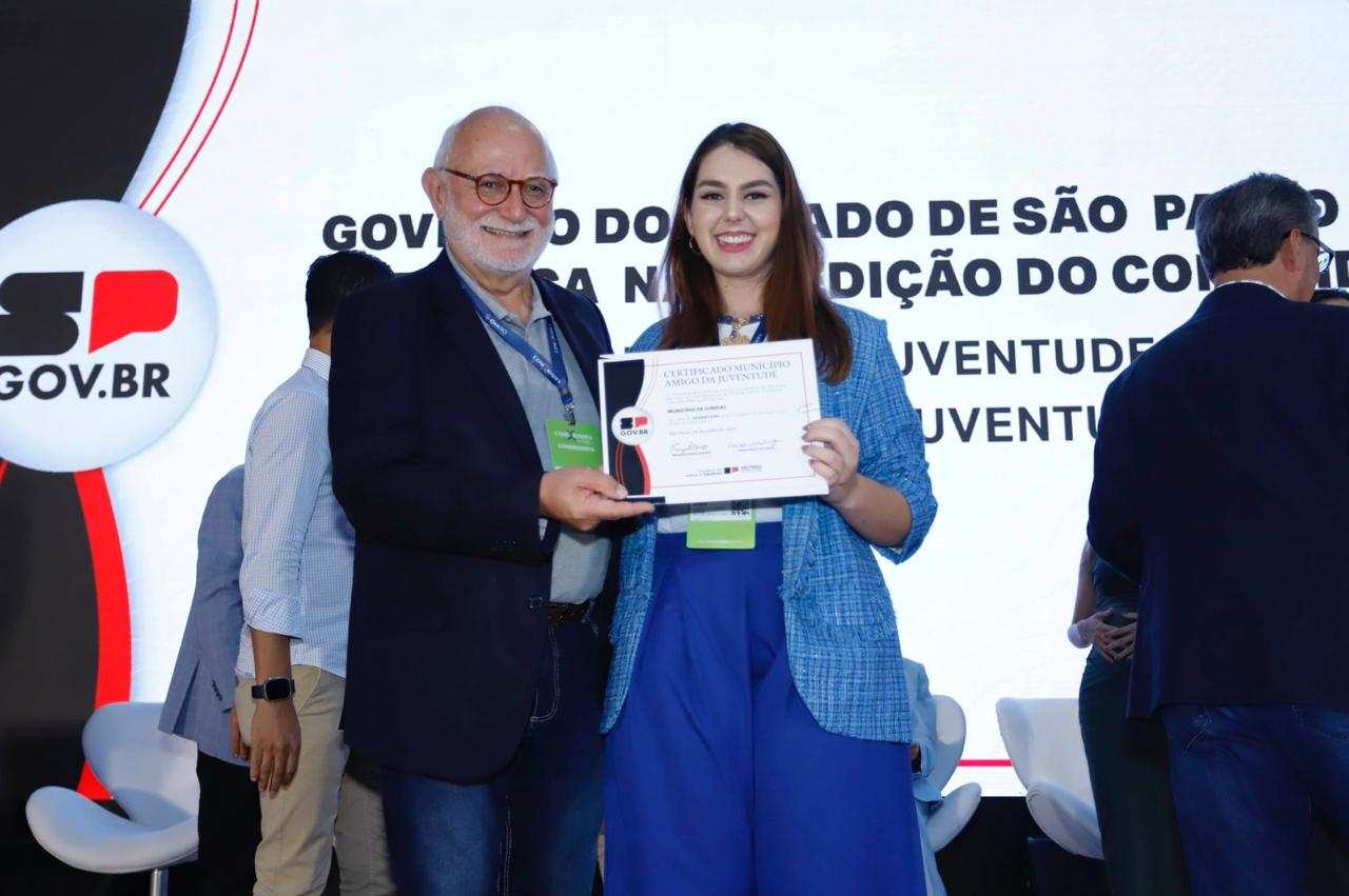 Homem idoso e mulher jovem seguram certificado de Certificação de Município Amigo da Juventude em evento do Governo do Estado de São Paulo, com logo e texto ao fundo.